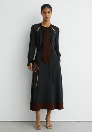 Ženska v črni midi obleki z rjavimi detajli, ki drži rjavo torbico z zlato verižico, stoji ob belem zidu.