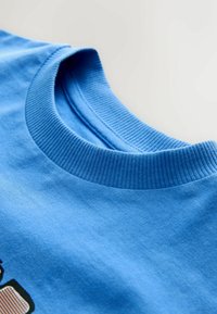 Kék pamut póló bordás kerek nyakkivágással, sima textúrával, és enyhén elhalványult nyomtatott részlettel az elülső részen.