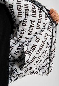 Jachetă captusită cu un model de text alb și negru, cu închidere cu fermoar și buzunar lateral, textură netedă și un design modern.