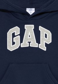 Námornícky modrá mikina s predným vreckom, so širokým šedým vyšitým nápisom "GAP". Vyrobená z mäkkej zmesi bavlny.
