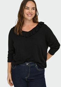 Nasmejana žena s dugom smešnom kosom koja nosi crni pulover s volanom na izrezu i tamnoplave traperice, nasuprot jednostavne svijetle pozadine.