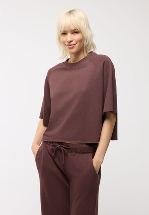 Giovane donna con capelli biondi corti che indossa una maglia marrone morbida a maniche corte e pantaloni abbinati con coulisse, mani in tasca, sorriso leggero.