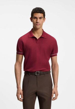 Mann trägt ein bordeauxfarbenes kurzärmeliges Strickpolo und braune Hose mit schwarzem Gürtel, steht vor weißem Hintergrund.