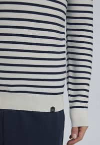 Pull en maille rayé crème et marine, avec des poignets et un ourlet côtelés, comportant une petite étiquette de marque en bas de l'ourlet.