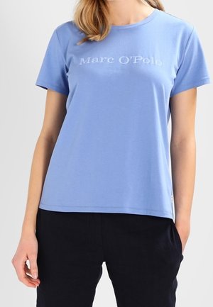 Vrouw draagt een lichtblauw T-shirt met korte mouwen met de tekst "Marc O’Polo" en zwarte broek, staand met één hand in de zak.