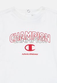 Λευκό πουκάμισο Champion με κόκκινο και γκρι περίγραμμα κειμένου "CHAMPION", κόκκινο λογότυπο "C" και κείμενο "Authentic Athleticwear" από κάτω.