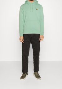 Hombre con una sudadera verde claro con bolsillo frontal, pantalones negros y zapatillas verde oliva, de pie contra un fondo blanco liso.