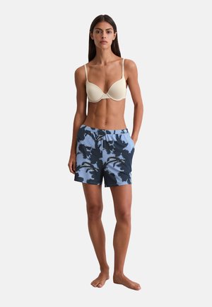Femme debout pieds nus, portant un soutien-gorge beige et un short à motifs floraux bleus, les mains dans les poches, devant un fond blanc.