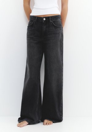 Persoon draagt een zwarte, wijde, hooggetailleerde jeans en een wit mouwloos topje, staat blootsvoets tegen een witte achtergrond.