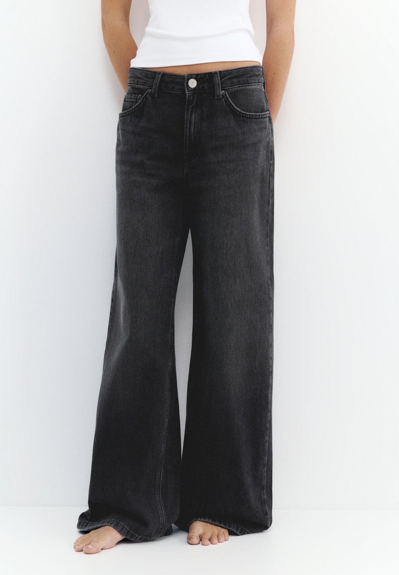 Persoană purtând blugi negri cu talie înaltă și picioare largi și un top alb fără mâneci, stând desculță pe un fundal alb.