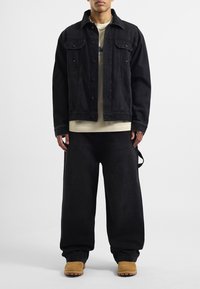 Zwarte denimjack met drukknoopjes, gecombineerd met een beige shirt en wijde zwarte broek. Bruine laarzen maken de outfit compleet.