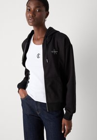 Black zip-up hoodie with front pockets and drawstring, featuring a logo on the chest. Worn over a white tank top and dark jeans.