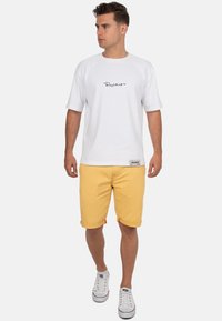 Witte katoenen t-shirt met zwart logo, gele opgerolde shorts en witte sneakers met rode accenten. Eenvoudig, casual outfitontwerp.