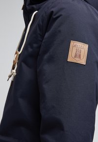 Marinblå jacka med strukturerad yta, prydd med en framträdande tanfärgad läderlapp med en logotyp, vita dragsko och träknappar.