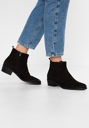 Person trägt blaue Denim-Jeans und schwarze Wildleder-Stiefeletten mit niedrigem Blockabsatz und seitlichen Reißverschlüssen.