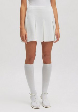 Woman wearing a white pleated mini skirt, knee-high white socks, and white strappy heels, standing on a plain light background.