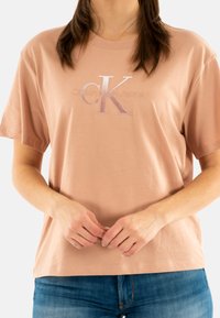 Camiseta de algodón de color rosa claro con mangas cortas, que presenta un logo bordado de "CK" y el texto "Calvin Klein Jeans" en un acabado brillante.