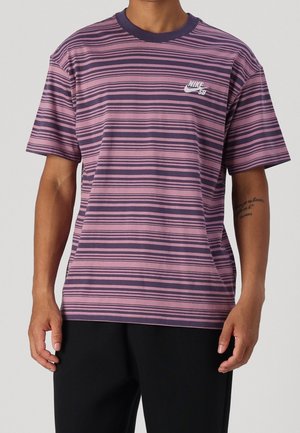 Mężczyzna ubrany w fioletowo-granatowy pasiasty t-shirt Nike SB z krótkim rękawem i okrągłym dekoltem oraz czarne spodnie, pokazujący mały tatuaż na ramieniu.