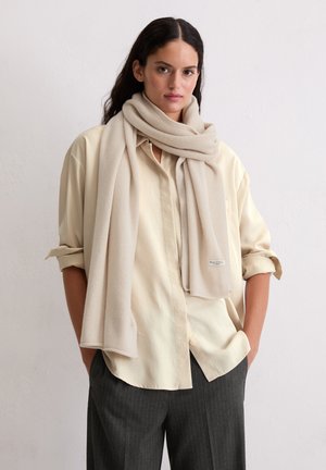 Young woman wearing a beige oversized shirt, light beige scarf wrapped around neck, and dark pinstripe trousers, standing with hands in pockets.