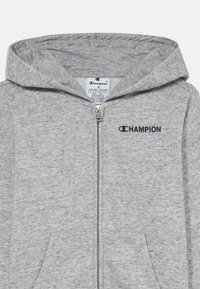Sudadera con cremallera de color gris claro, con capucha, bolsillos frontales y logo negro de Champion en el lado izquierdo del pecho, encima de la cremallera.