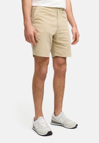 Beige shorts med flat front, belteheft, side lommer og en avslappet passform. Kombinert med lysegrå sportsko med svarte detaljer.