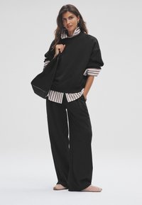 Must oversized dressipluus, triibuline särk all, mustad laiade säärega püksid ja pehmed roosad kortsud. Aksessuaaridena must õlakott.