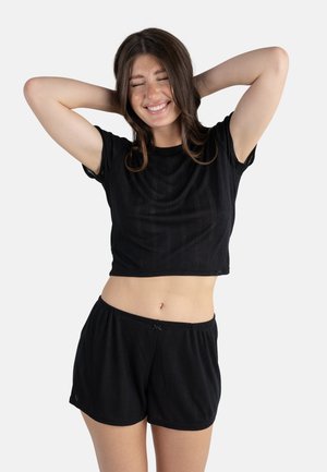 Femme aux longs cheveux bruns en crop top noir à manches courtes et short, souriant les yeux fermés avec les mains derrière la tête.