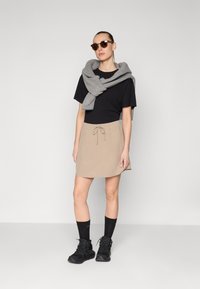 Falda beige con cordón, camiseta negra, bufanda gris, gafas de sol negras y zapatos de senderismo negros con calcetines oscuros. Diseño simple, aspecto casual.