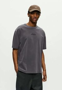 Νεαρός άνδρας που φοράει ένα ξεθωριασμένο γκρι φαρδύ μπλουζάκι με μικρό κείμενο στο στήθος, σκούρα παντελόνια και μπεζ καπέλο, σε απλό ανοιχτό φόντο.