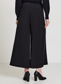 Donna che indossa pantaloni neri a gamba larga e cropped, un top nero a maniche lunghe e scarpe con tacco a blocco nere, vista da dietro.