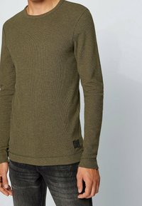 oliwkowa koszula z długim rękawem o prążkowanej teksturze, okrągłym dekolcie i małym czarnym detalu z metką u dołu, w połączeniu z ciemnymi dżinsami.