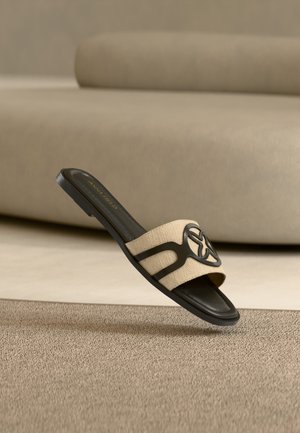 Sandale plate noire et beige à enfiler, à bout ouvert avec un emblème d'étoile sur la bride en tissu, posée sur un tapis texturé devant un canapé beige.