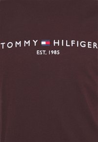 Mørkebrun t-shirt med hvid broderet tekst "TOMMY HILFIGER" og "EST. 1985," samt en lille blå, rød og hvid logoakkord.