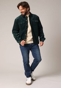 Grüne Cordjacke mit zwei Brusttaschen, getragen über einem leichten cremefarbenen Pullover, kombiniert mit blauen Jeans und weißen Sneakers.