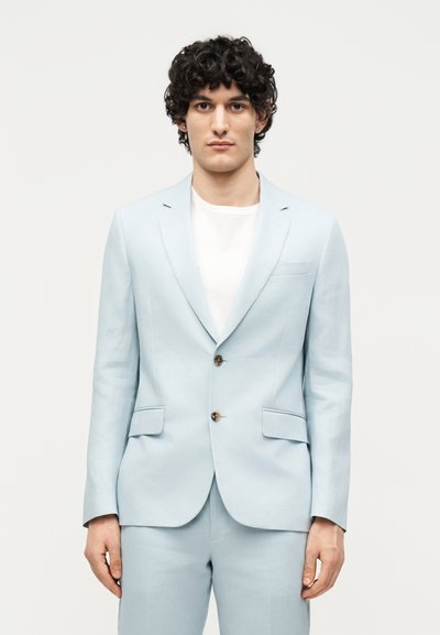 Άνδρας με σγουρά σκούρα μαλλιά φοράει ανοιχτά μπλε σακάκι με δύο κουμπιά και παντελόνι που ταιριάζει πάνω από λευκό πουκάμισο, στέκεται μπροστά από απλό φόντο.