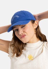 Blue cotton cap with curved brim, embroidered "ROXY 1990" on front. Model wears a cream top with yellow floral embroidery, showing arm tattoo.