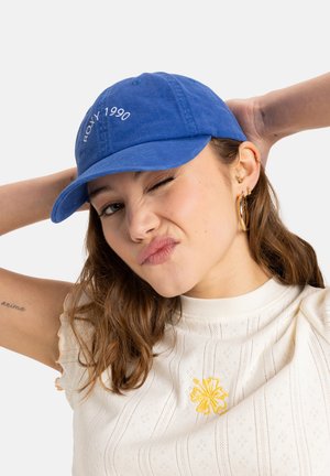 Blå bomuldske med buet skygge, broderet "ROXY 1990" foran. Modellen har en cremefarvet top på med gule blomsterbroderier og viser en arm tatovering.