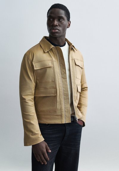 Veste fauve ajustée avec de grandes poches avant et une fermeture éclair. Portée sur un pull crème. Un jean foncé complète la tenue. Texture de tissu lisse.