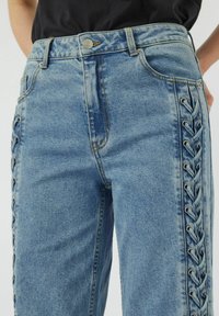 Jeans de mezclilla azul de cintura alta con un diseño de lado con cordones y ojetes metálicos, cinco bolsillos y detalles de costura visibles.