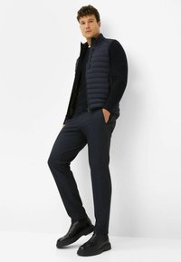 Schwarze Steppweste über einem marineblauen Langarmshirt, kombiniert mit dunkel gemusterten Hosen und schwarzen Ankle Boots. Einfaches, modernes Design, figurbetonter Schnitt.