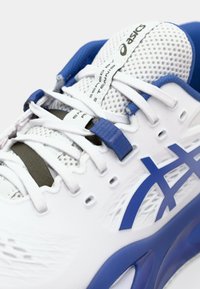 Chaussure de sport blanche avec des accents bleus, en mesh et en synthétique, dotée de lacets, d'une étiquette de logo et de détails texturés sur la semelle.