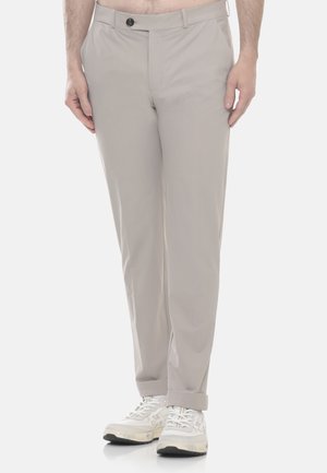 Uomo che indossa pantaloni beige chiaro e scarpe da ginnastica bianche, in piedi con le mani rilassate lungo i fianchi su uno sfondo semplice.