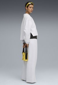 Sudadera blanca recortada con acentos negros, combinada con pantalones anchos blancos. Sosteniendo un bolso pequeño amarillo con correa negra y diseño decorativo.