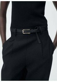 Cintura in pelle nera con fibbia dorata, indossata sopra pantaloni neri a vita alta e sagomati con tasche laterali e una maglietta nera strutturata.