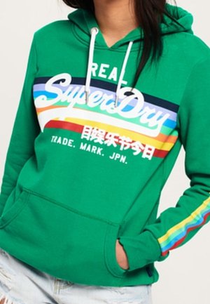 Green hoodie with white drawstrings, rainbow stripes, "Real Superdry" text, Japanese characters, and a front pocket, worn with light ripped jeans.