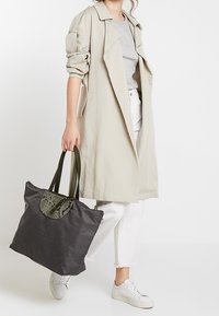 Szara torba typu tote z teksturowanym czarnym akcentem, wyposażona w dwie solidne zielone rączki. Modelka ma na sobie jasnobeżowy płaszcz i białe spodnie oraz szary top.