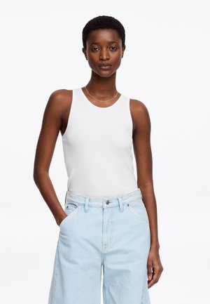 Femme aux cheveux courts portant un haut blanc sans manches et un jean taille haute bleu clair, debout avec une main dans la poche.