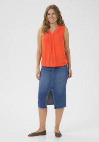 Haut orange sans manches avec un détail de bouton et un décolleté en V profond, associé à une jupe en denim avec une fente sur le devant, et des chaussures plates marron.