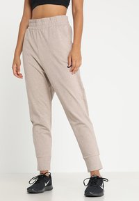 Pantalones de chándal color beige claro con una textura suave, cinturilla elástica y tobillos ajustados; combinados con zapatillas deportivas negras que tienen un logo blanco.