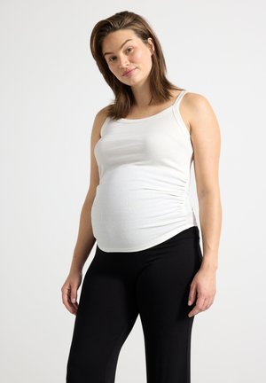 Gravid kvinde iført en hvid ærmeløs top og sorte leggings, stående mod en ensfarvet lys baggrund og ser på kameraet.